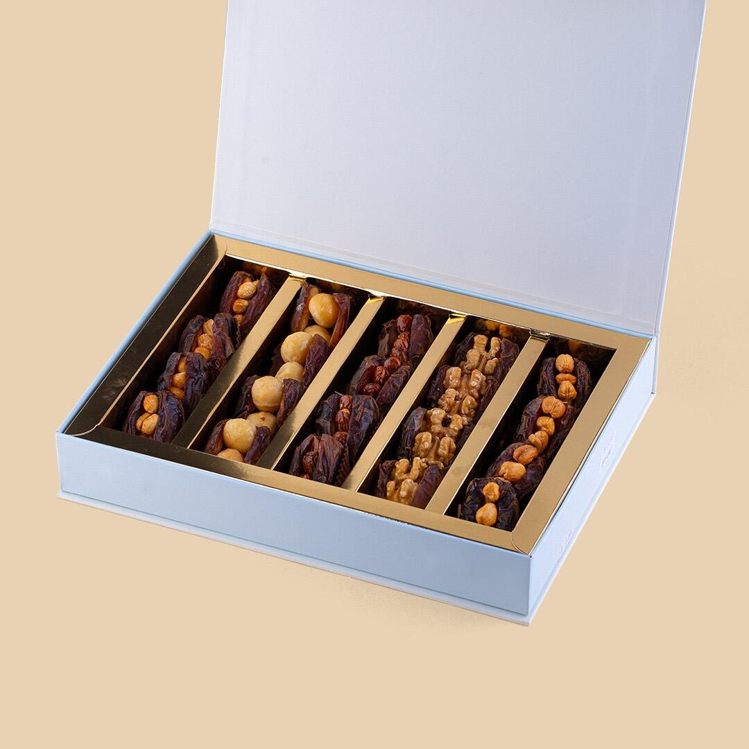 An open white gift box featuring five rows of jumbo stuffed dates topped with assorted nuts like walnuts, almonds, and macadamias on a light beige background