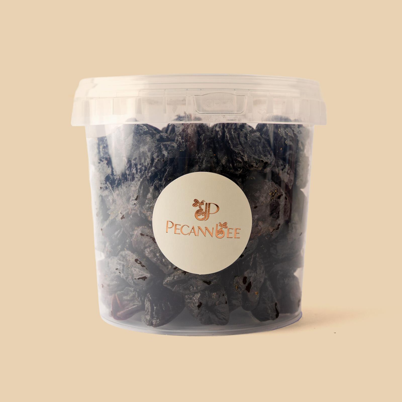 A clear plastic tub filled with dark, dried seedless prunes, featuring a white circular label with the gold Pecanbee logo, set against a solid beige background