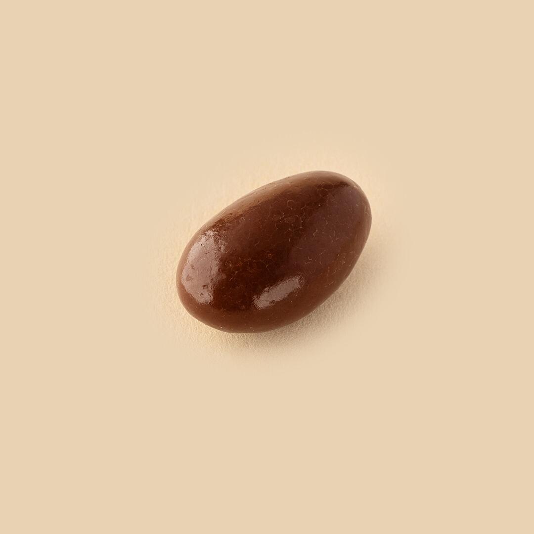 A single, smooth milk chocolate-coated almond dragee isolated on a solid light beige background, viewed from a slightly elevated angle.
