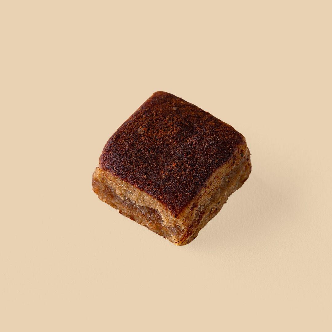 A single square-shaped Rangina treat with a dark, spiced crust and a dense date-paste filling, presented at an angle against a solid neutral beige background