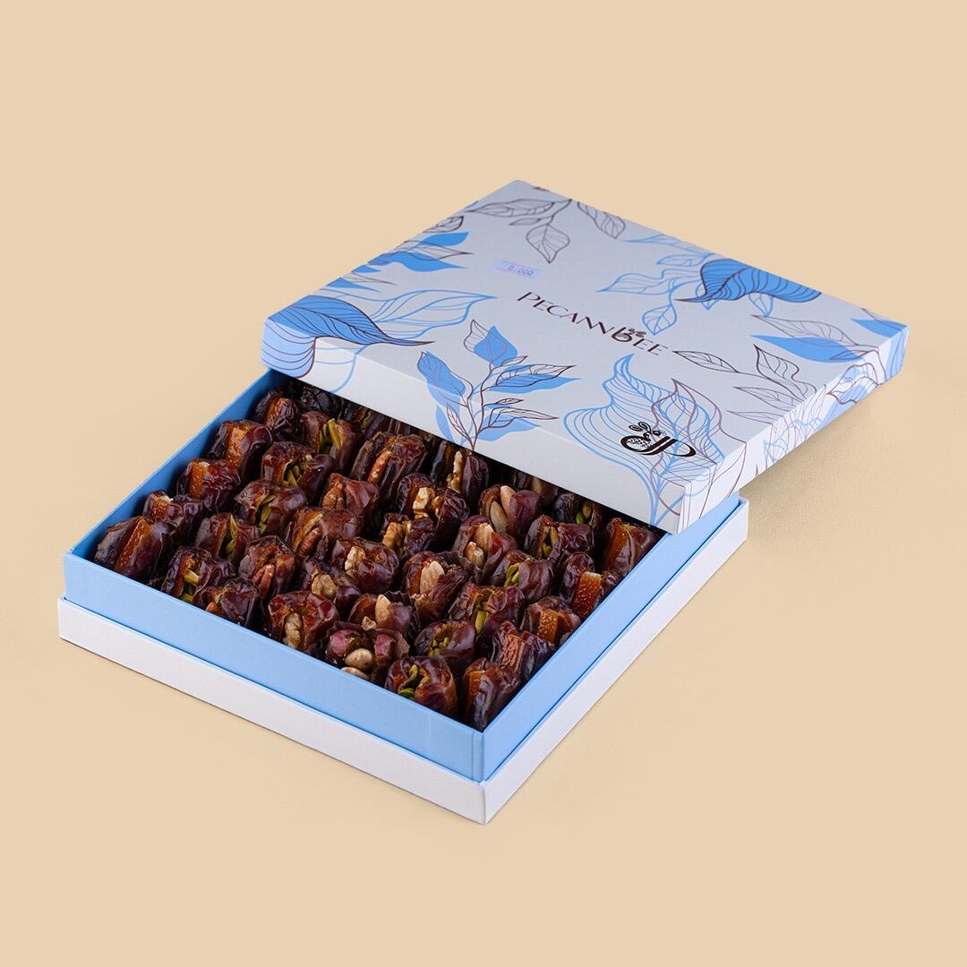 Open gift box of Khudri dates stuffed with mixed nuts, featuring a blue and white botanical patterned lid, shot at a high angle on a solid beige background