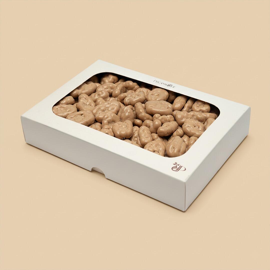 A white rectangular gift box with a clear window filled with light brown chocolate-coated pecan dragees, angled view against a solid beige background