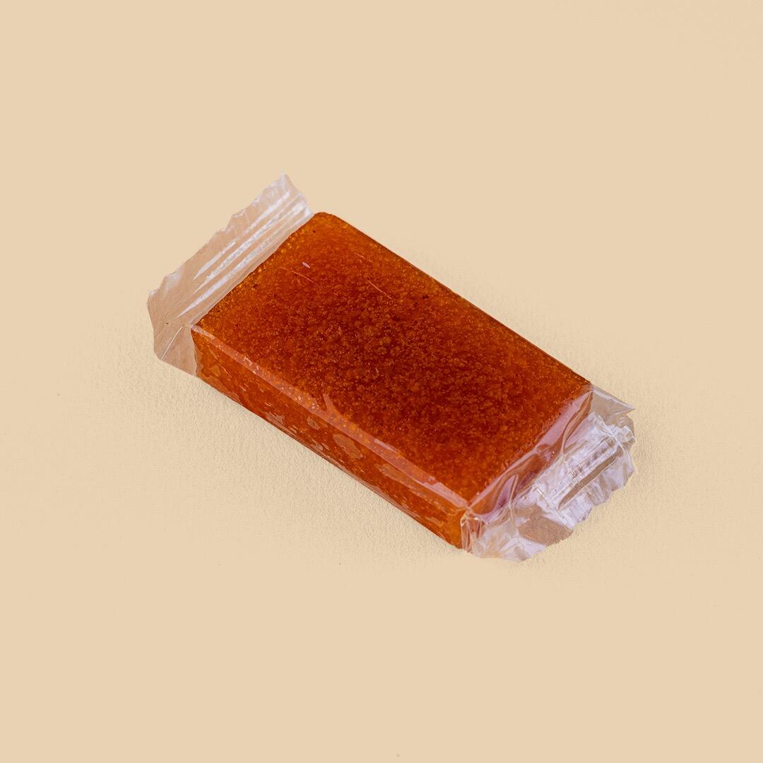 A rectangular, golden-orange apricot fruit leather bar individually wrapped in clear plastic, presented at a high angle against a plain light beige background