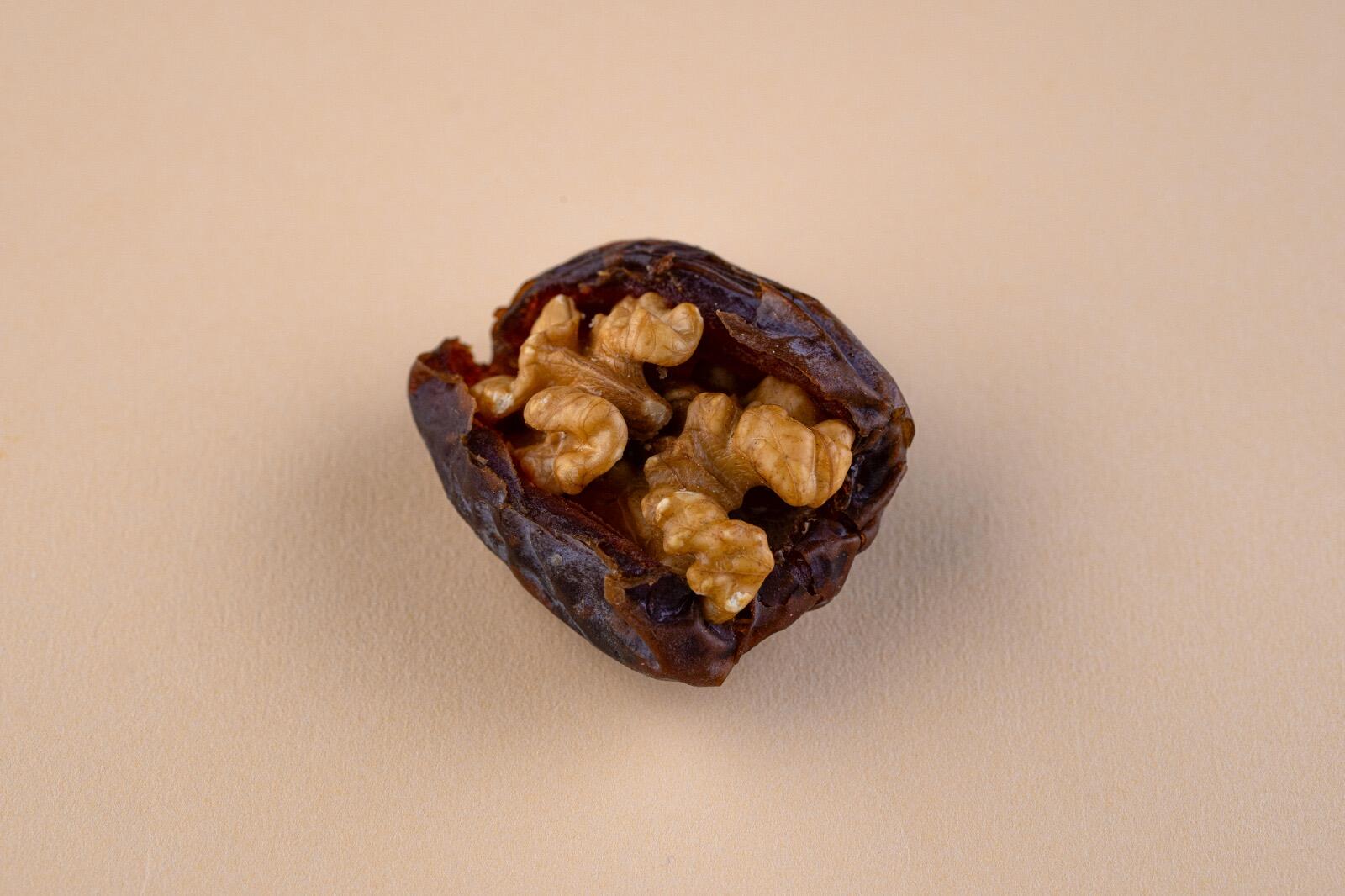 A close-up studio shot of a single jumbo Medjool date stuffed with a walnut half, centered against a solid light beige background, viewed from a slightly high a