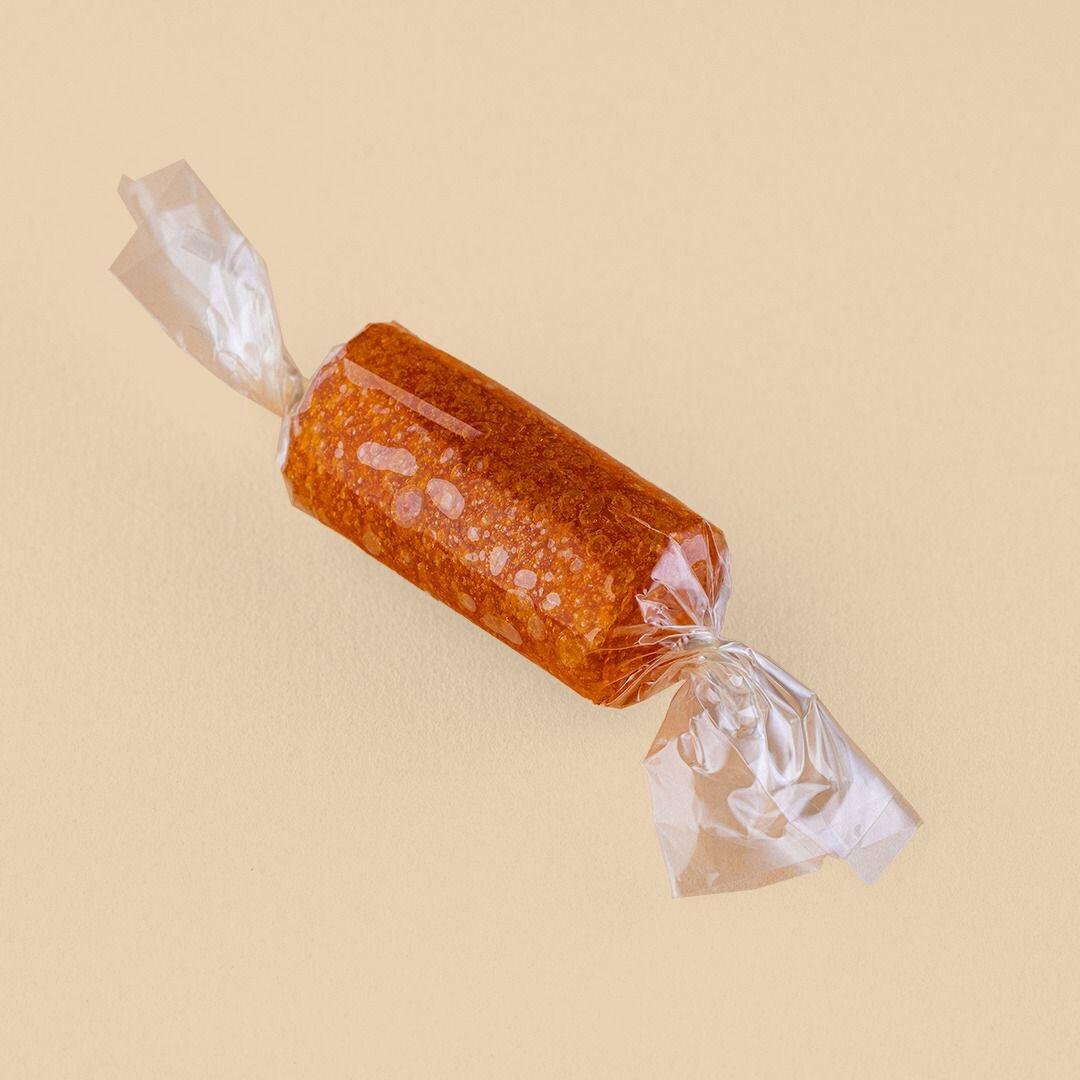 A single cylindrical piece of translucent orange dried apricot paste, or Qamar Al-Deen, wrapped in clear crinkled plastic against a solid beige background
