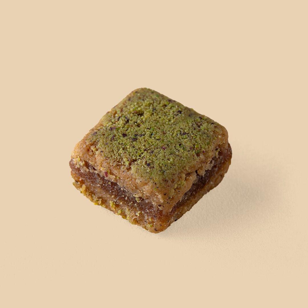 A single, square-shaped Rangina pastry topped with finely ground green pistachios, centered on a solid, light beige background.