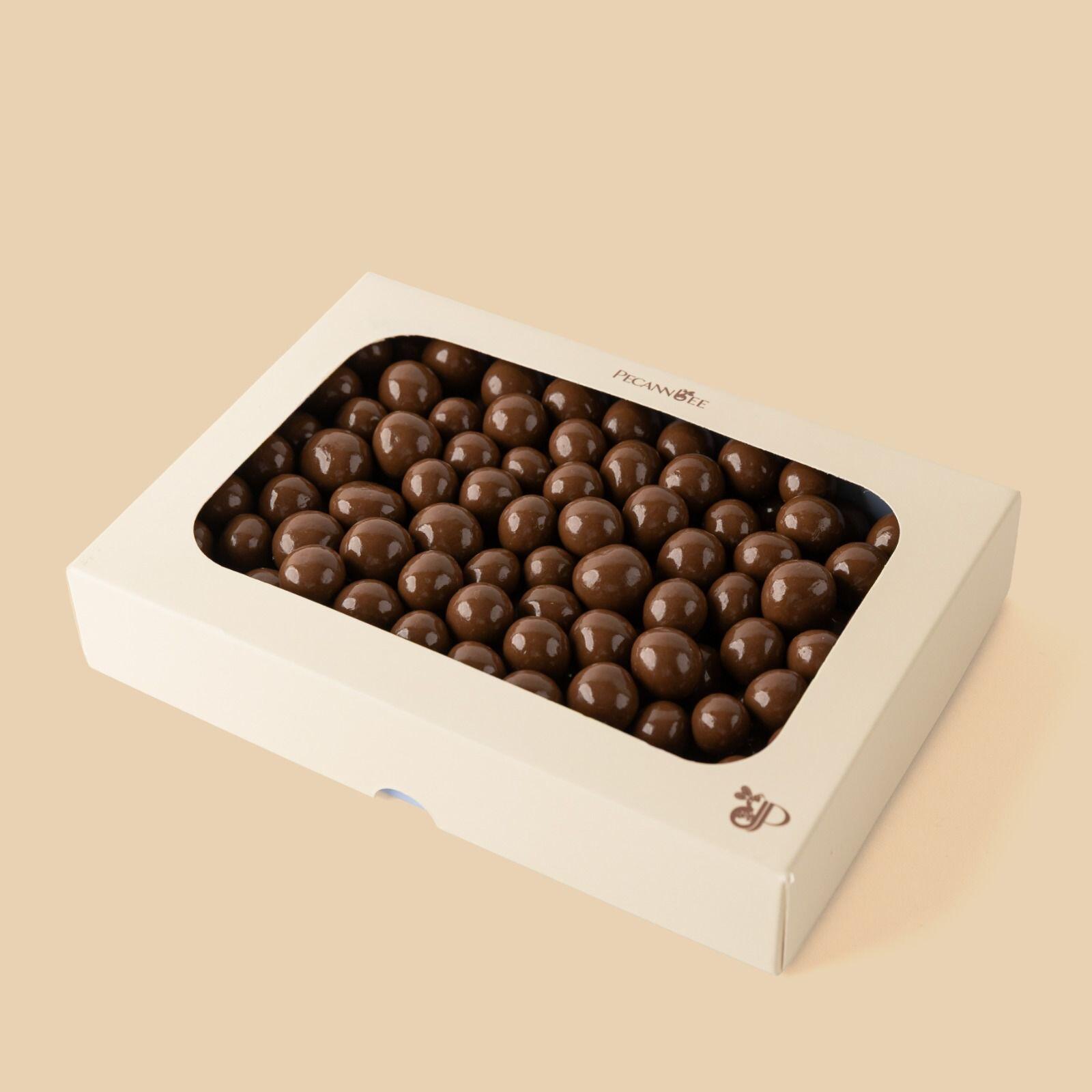 A white rectangular box with a clear window containing milk chocolate-coated hazelnuts, displayed at a high-angle shot against a solid beige background