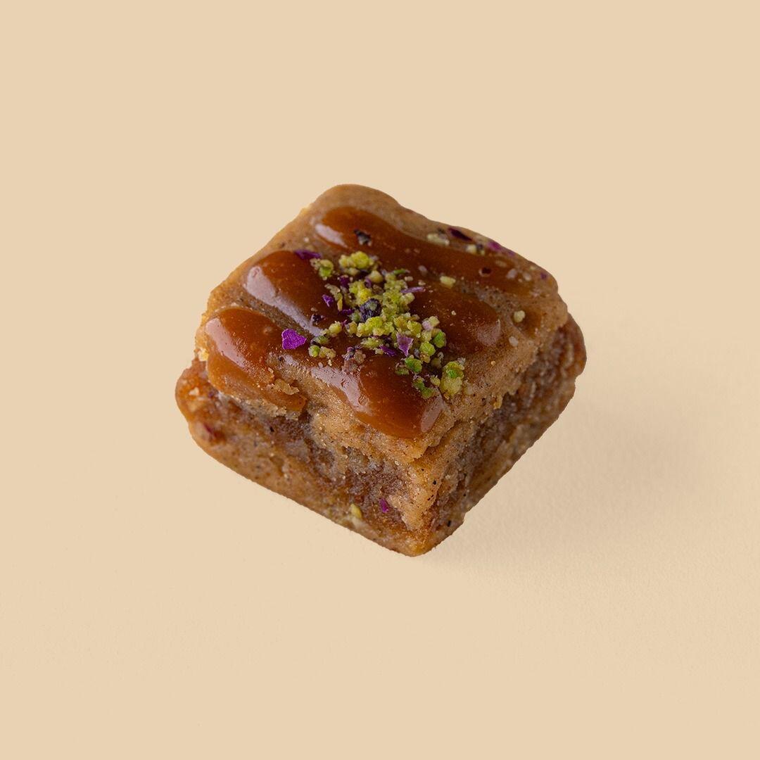A square piece of golden-brown Rangina dessert topped with a glossy caramel drizzle, crushed pistachios, and tiny purple flower petals against a beige backgroun