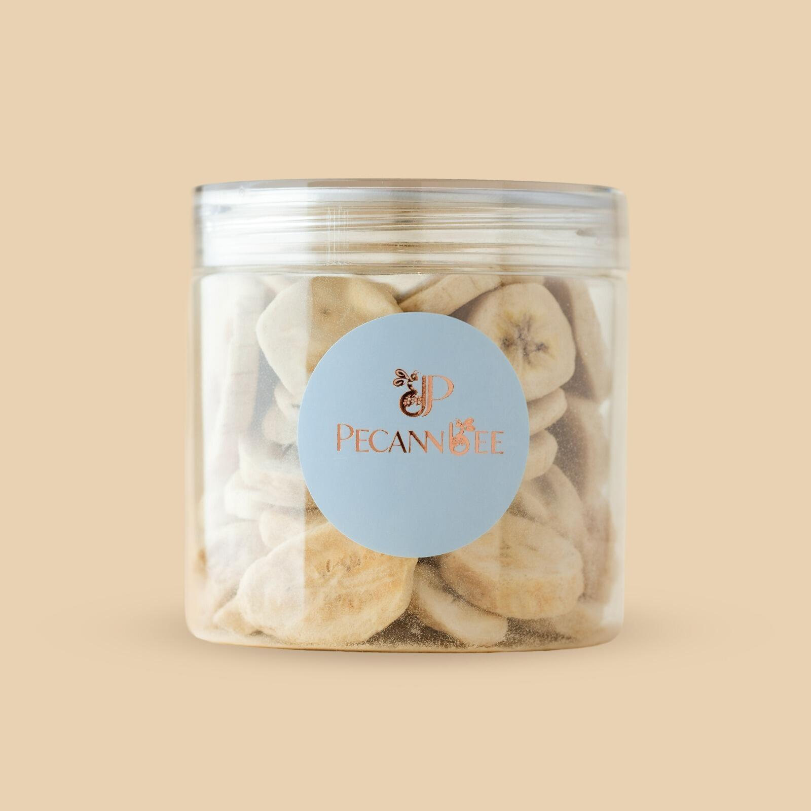 Clear plastic jar filled with freeze-dried banana slices, featuring a circular blue PecanBee brand label, set against a solid beige background