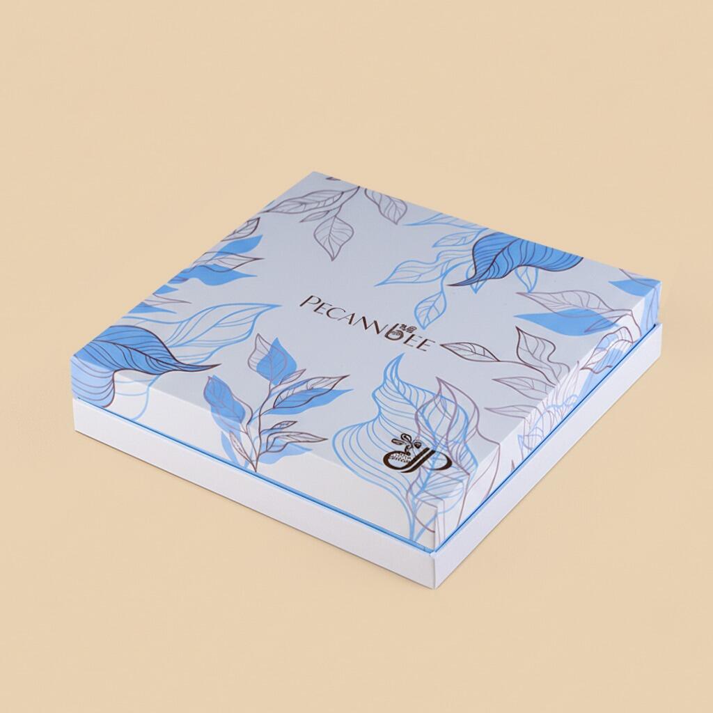 A high-angle studio shot of a square gift box with a light blue botanical leaf pattern and PecanBee branding, resting on a clean, solid beige background.