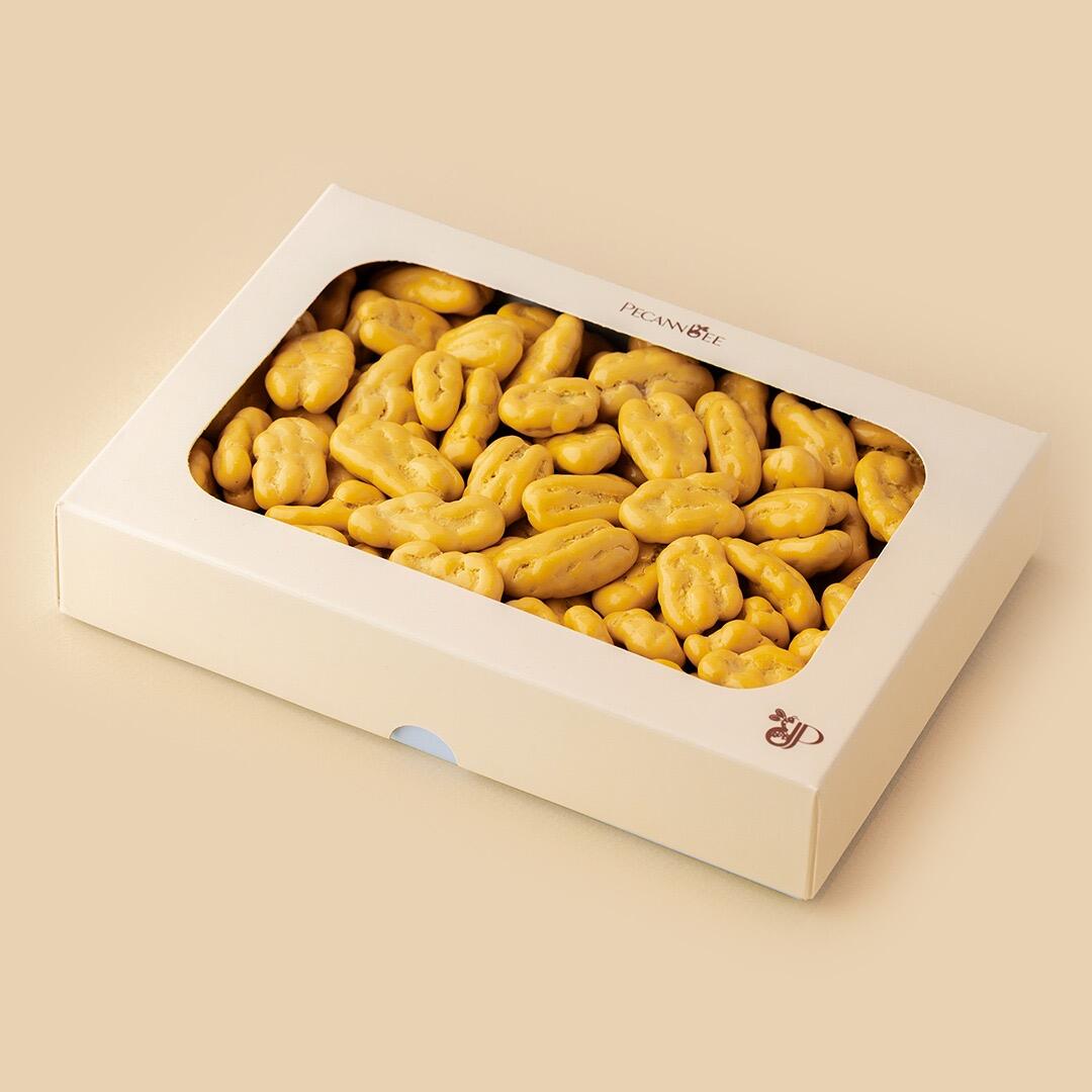 A rectangular white box with a clear window filled with yellow cheese cake-flavored chocolate-coated pecans, set against a solid beige background