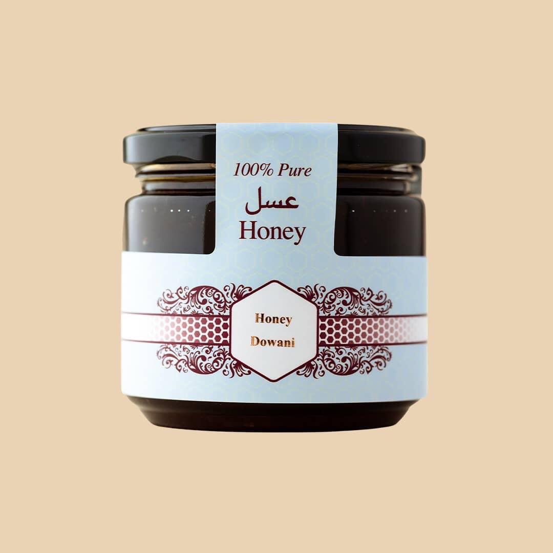 Small glass jar of dark liquid honey with a light blue label featuring Arabic and English text 100% Pure Honey and Dowani brand centered on a solid tan backgrou