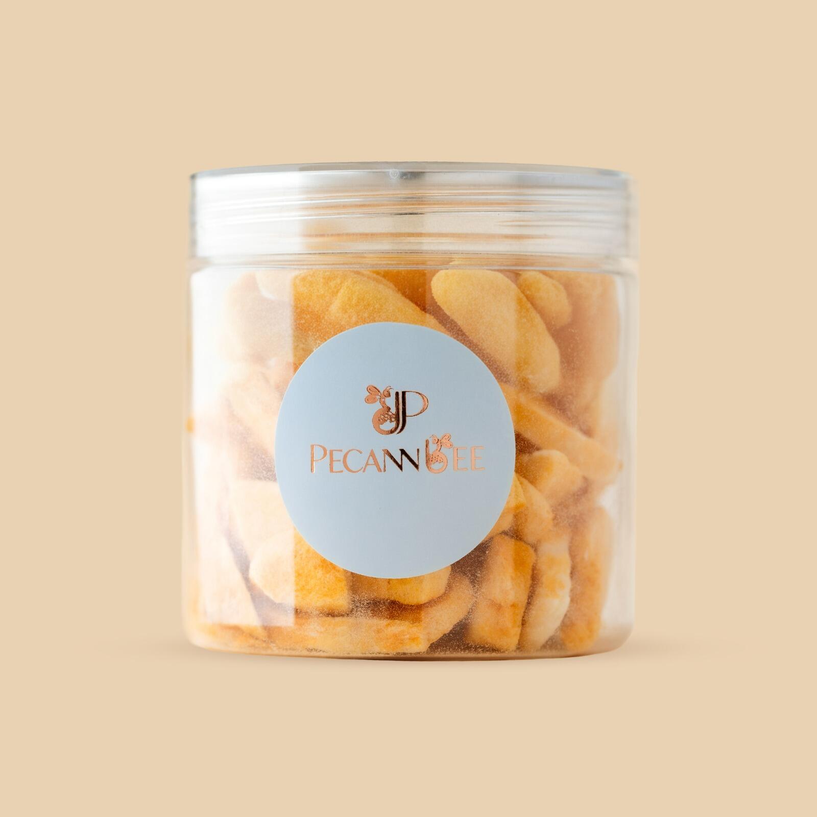 Clear cylindrical plastic jar filled with bright orange dried mango slices, featuring a round light blue logo label, centered against a solid beige background