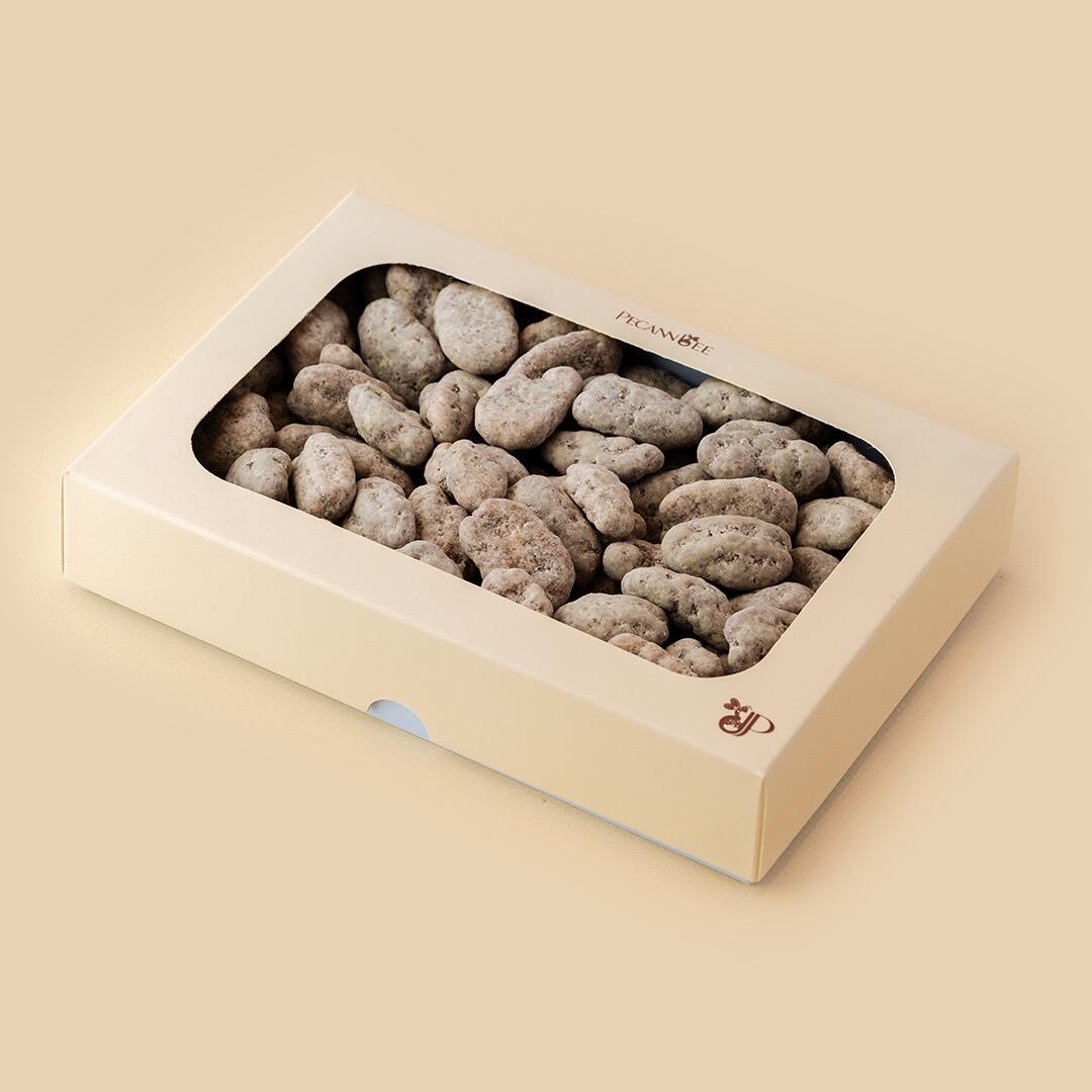 A rectangular beige gift box filled with chocolate-covered pecan dragees, angled from above against a matching light tan background, featuring a clear window to