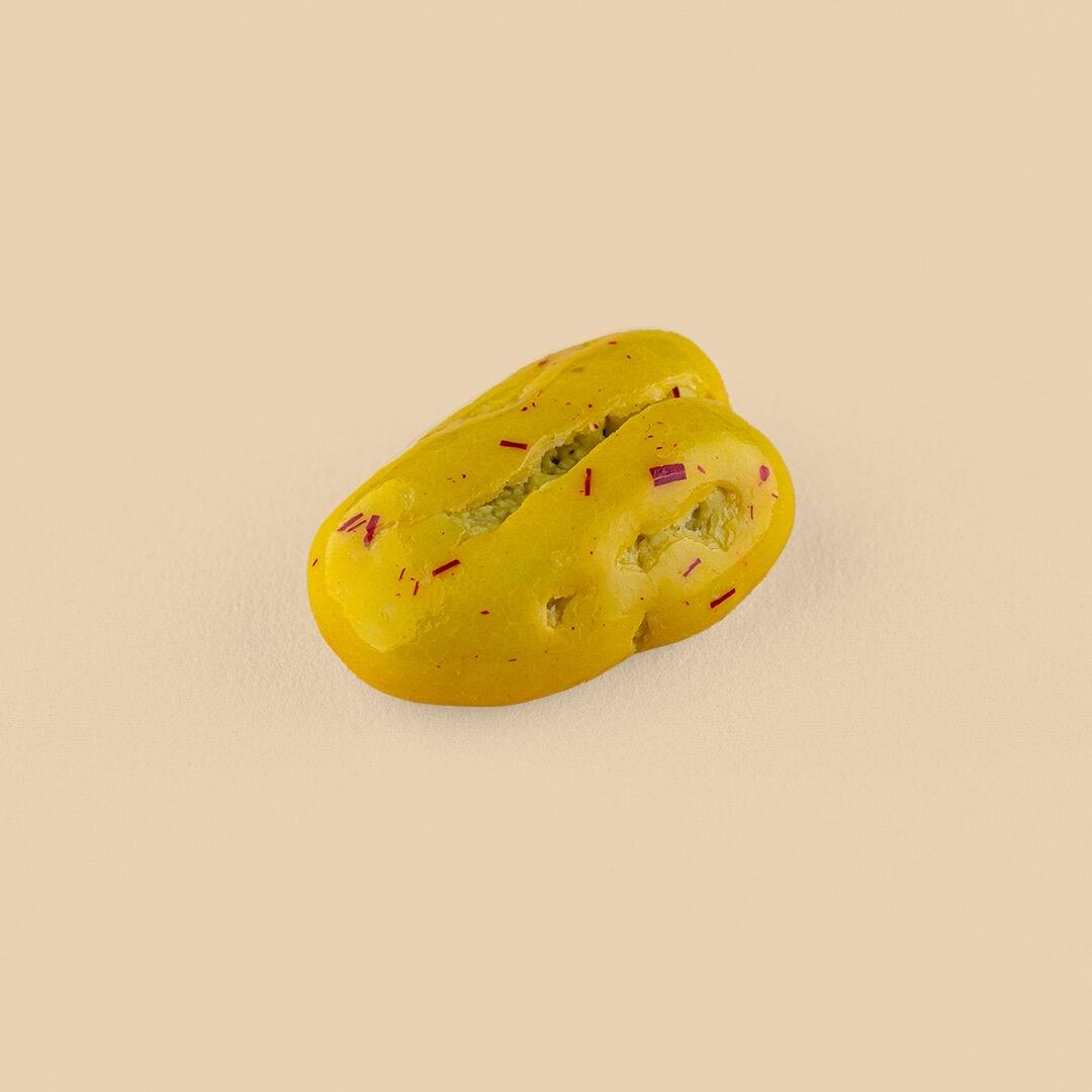 A single yellow, oval-shaped pecan dragée with a smooth, glossy saffron-infused coating and tiny red flecks, centered against a solid light beige background
