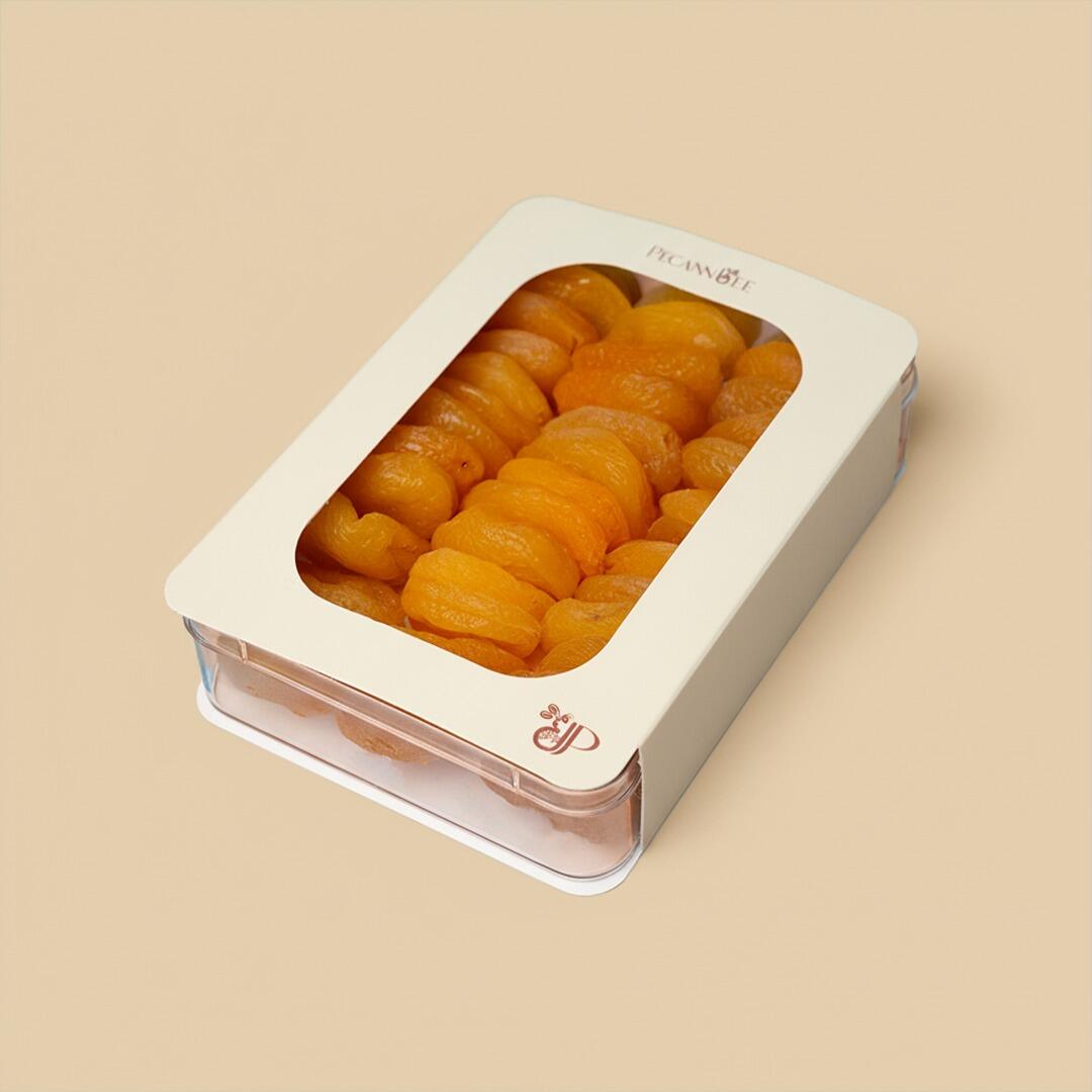 High-angle view of a clear plastic rectangular container filled with jumbo dried apricots, encased in a cream-colored sleeve with Pecanbee branding on a beige b