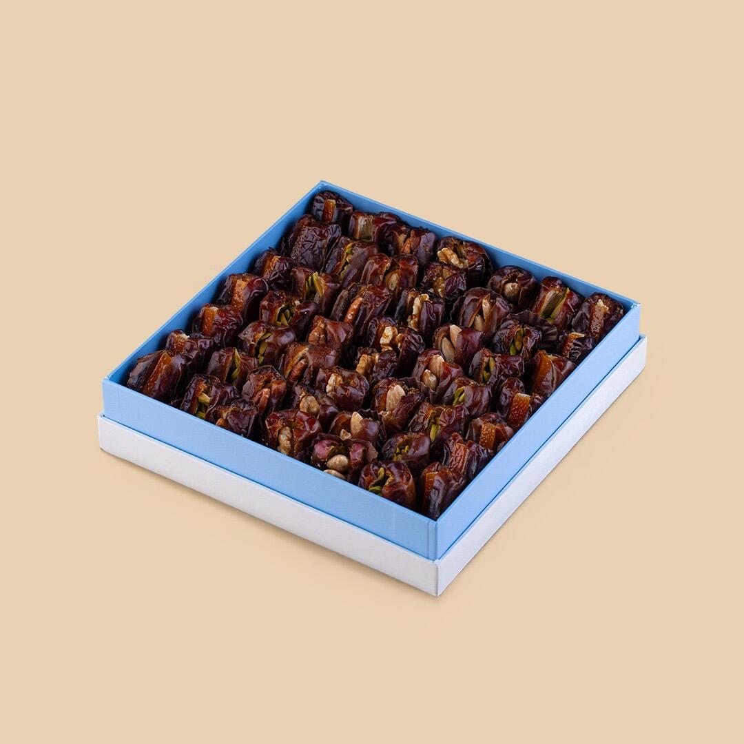 High-angle view of a square light blue box filled with Khudri dates stuffed with various nuts, set against a solid beige background