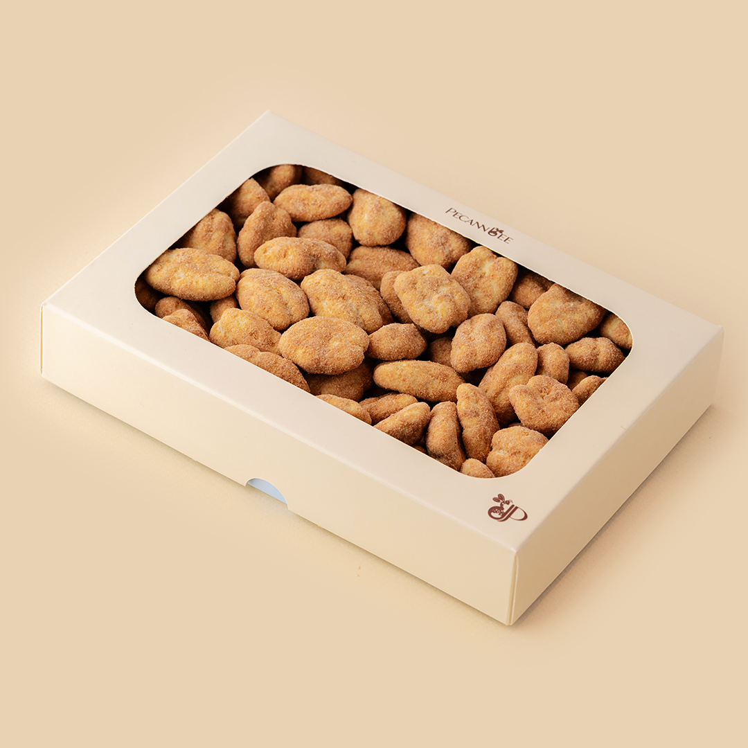 A rectangular beige box with a clear window filled with sugar-coated, light brown dragee pecans on a solid matching background, featuring Pecannbee branding