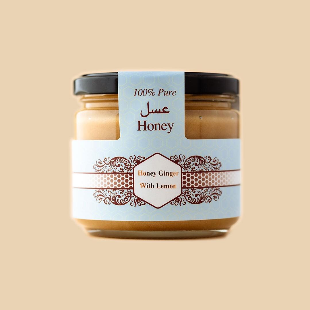 A glass jar of ginger honey with lemon, featuring a blue and white patterned label with Arabic and English text, centered against a solid light beige background