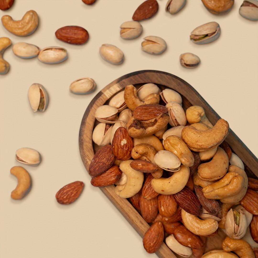 A rustic wooden bowl filled with a premium assortment of roasted almonds, cashews, and pistachios against a clean light beige background with scattered nuts nea
