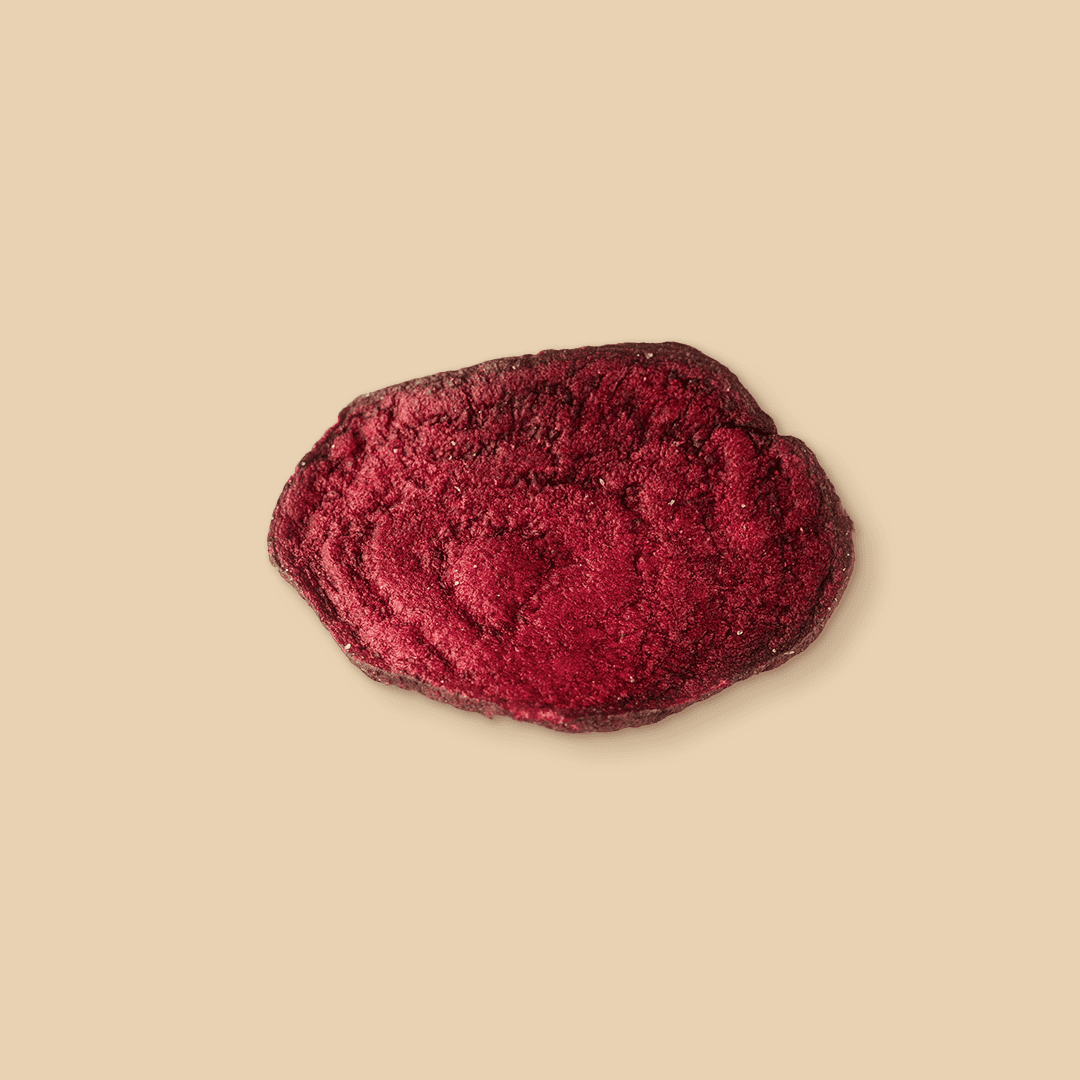 A single dehydrated beetroot slice with a deep red color and textured surface, shown from a top-down angle against a solid, light beige studio background