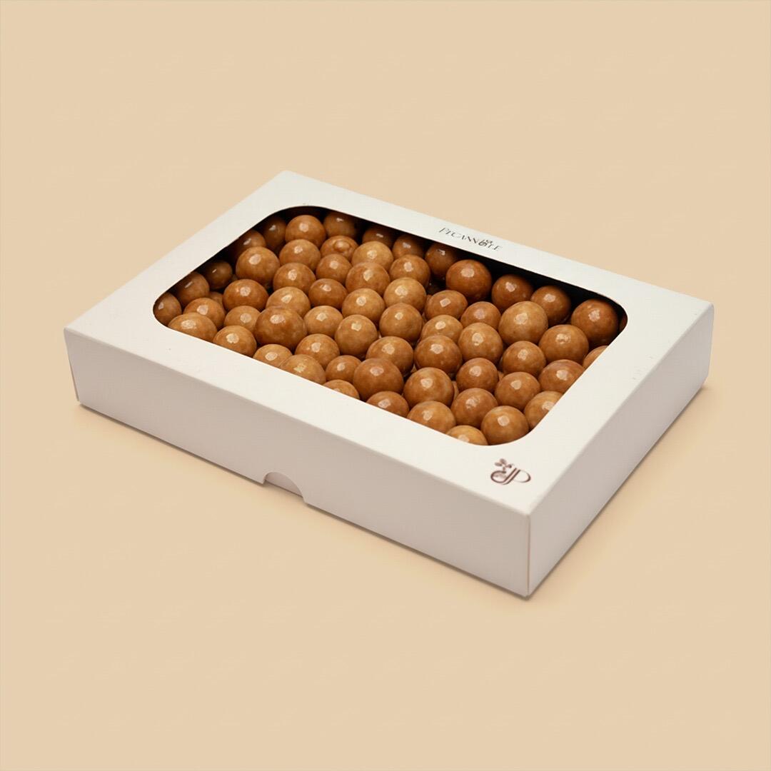 A rectangular white box with a window top filled with glossy, light brown caramel-coated hazelnut dragees, angled on a plain light beige background