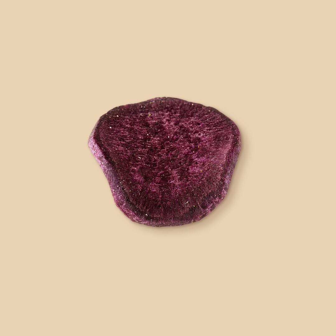 A single, round slice of dried, deep purple sweet potato seen from a top-down angle, isolated against a solid light beige background