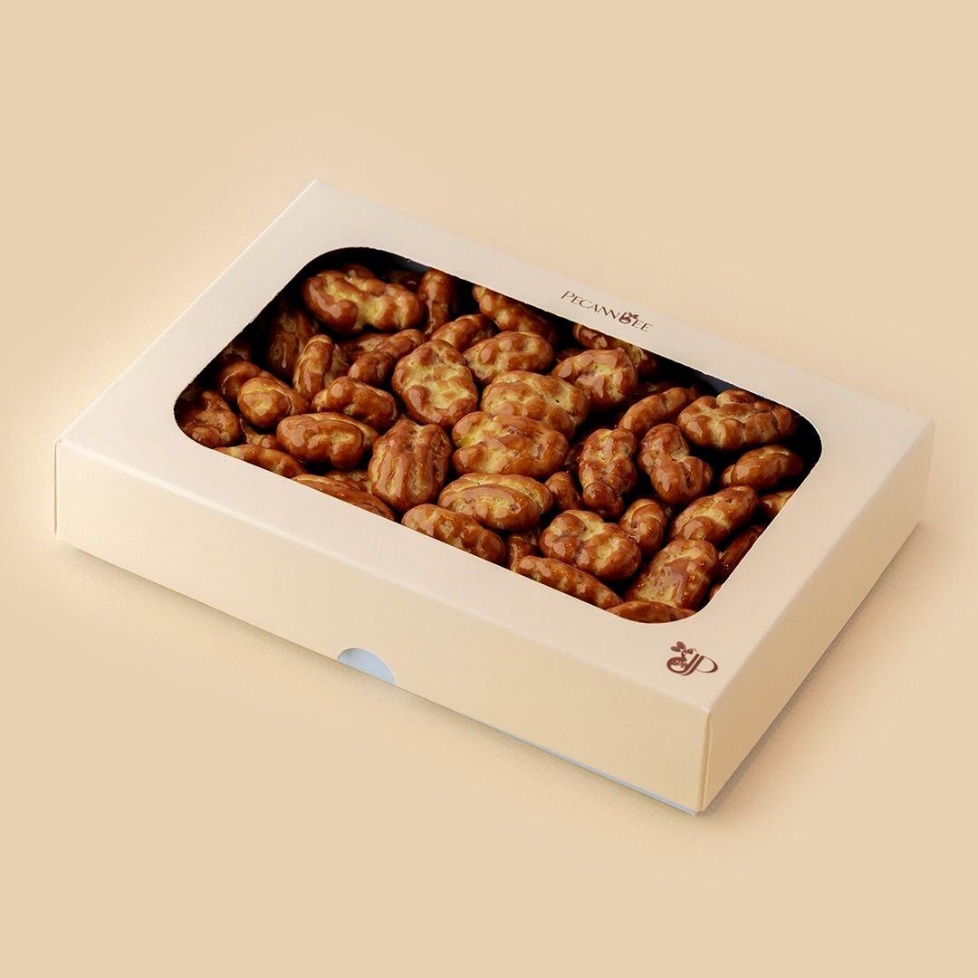 A high-angle view of a rectangular beige box with a transparent window filled with glossy, glazed pecans, featuring the PecanBee logo on a clean, light-colored 