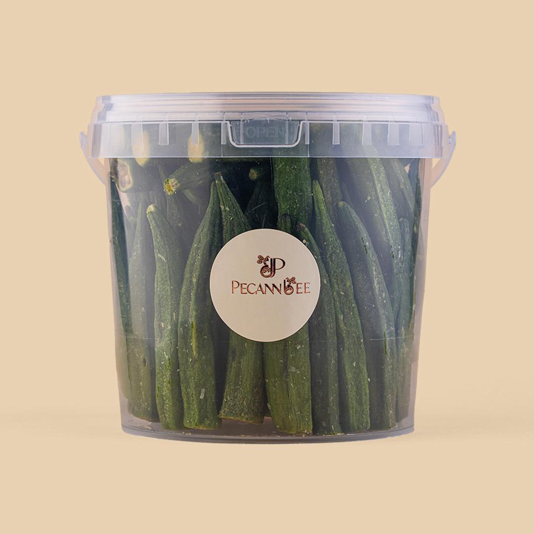 Clear plastic bucket filled with whole dried green okra pods, featuring a circular PecanBee brand label, set against a solid beige studio background