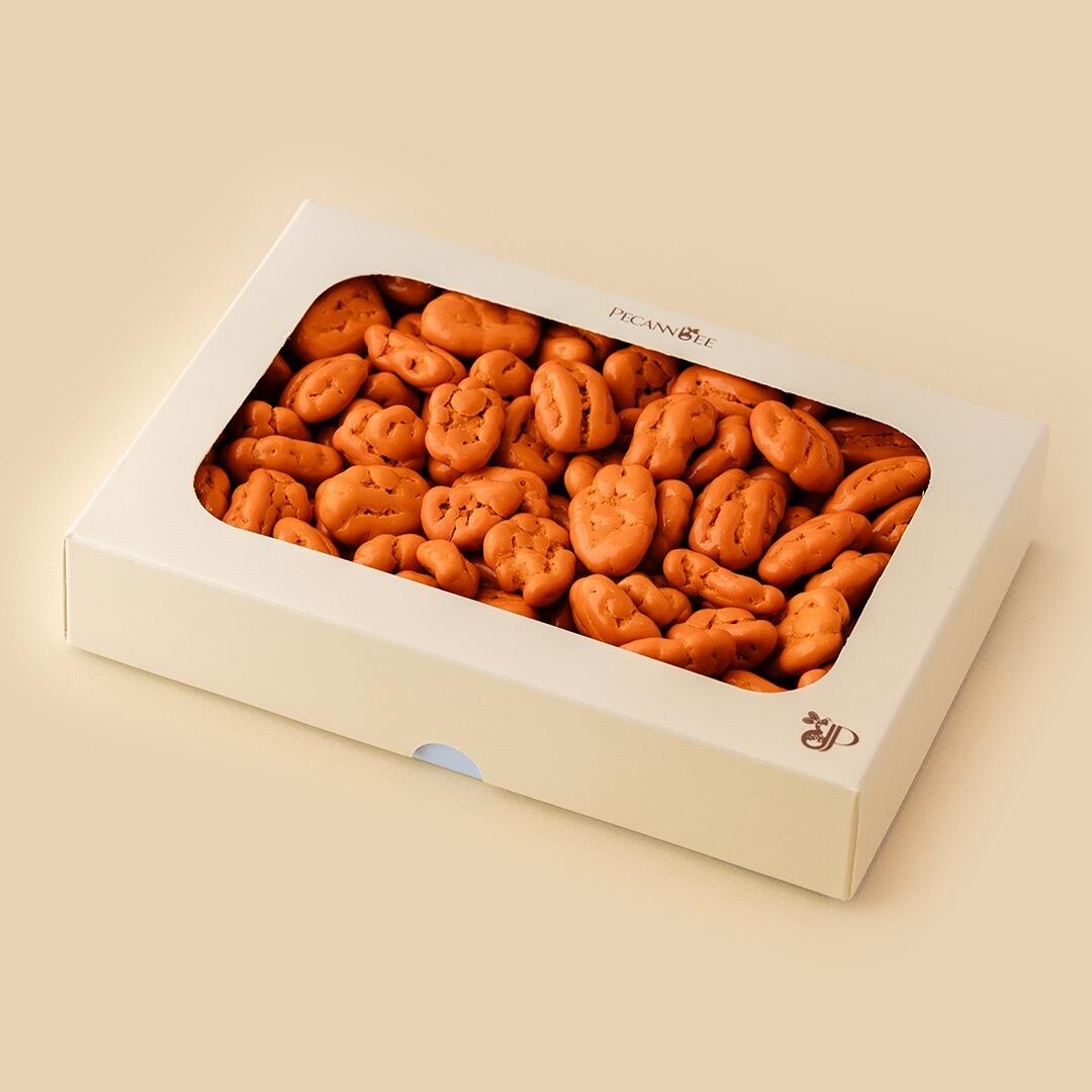A white rectangular gift box with a transparent window filled with bright orange, chocolate-coated pecan dragees, shot at an eye-level angle on a beige backgrou
