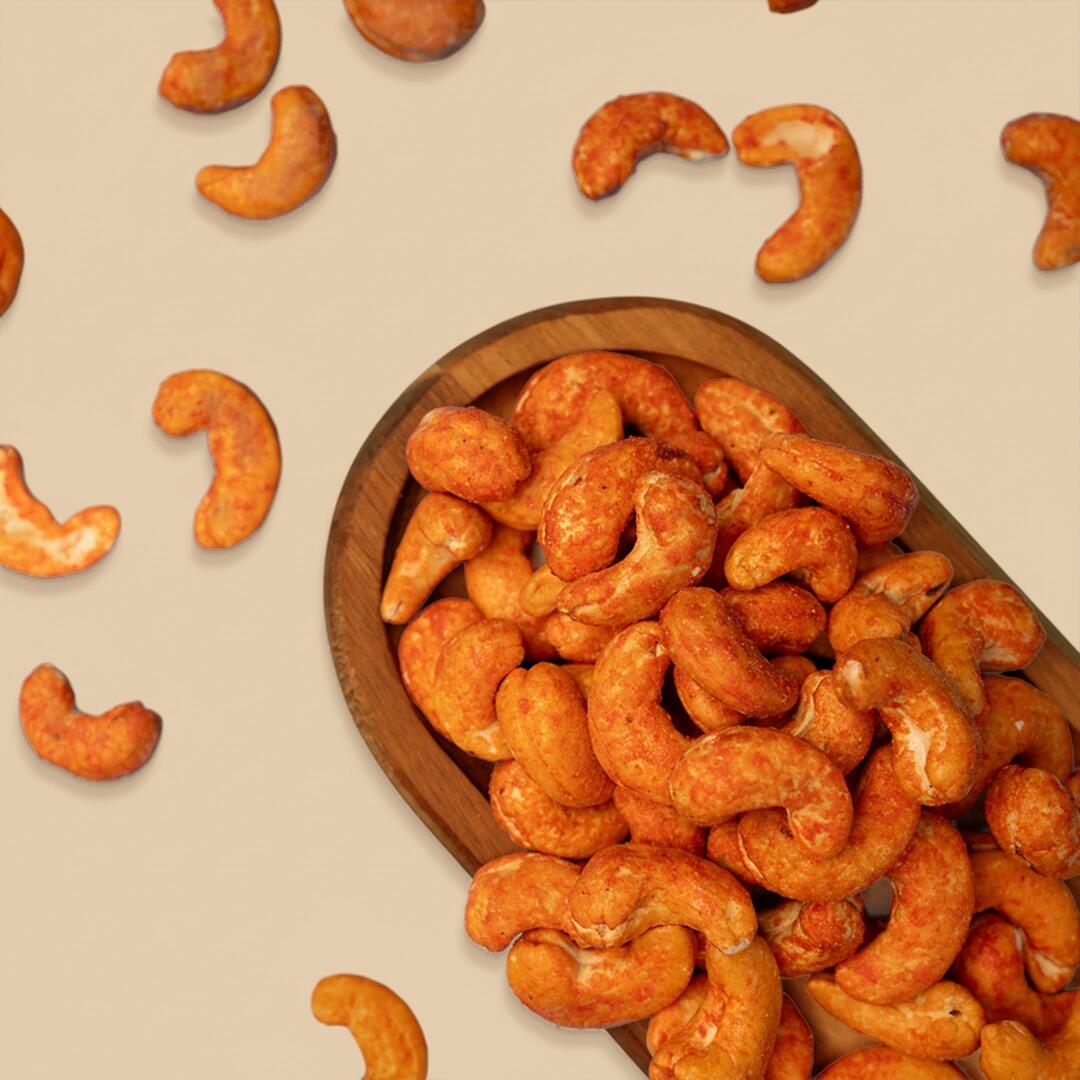Top-down view of a wooden oval bowl filled with bright orange, cheese-seasoned roasted cashews, with individual cashews scattered on a light beige background