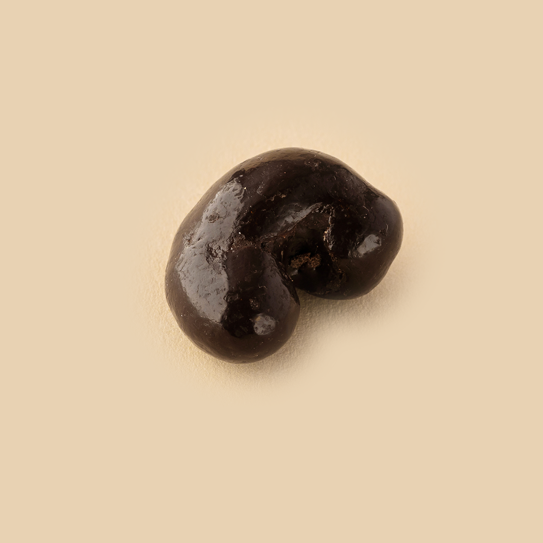 A single dark chocolate-covered cashew nut isolated on a solid light beige background viewed from a top-down angle