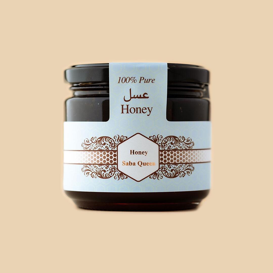 Small glass jar of Saba Queen honey with a light blue label featuring an ornate gold-tone border and Arabic text, centered against a neutral beige background