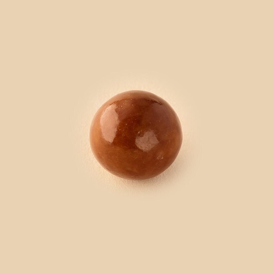 A single, round, glossy, caramel-colored hazelnut dragee centered against a smooth, solid beige background from a top-down view