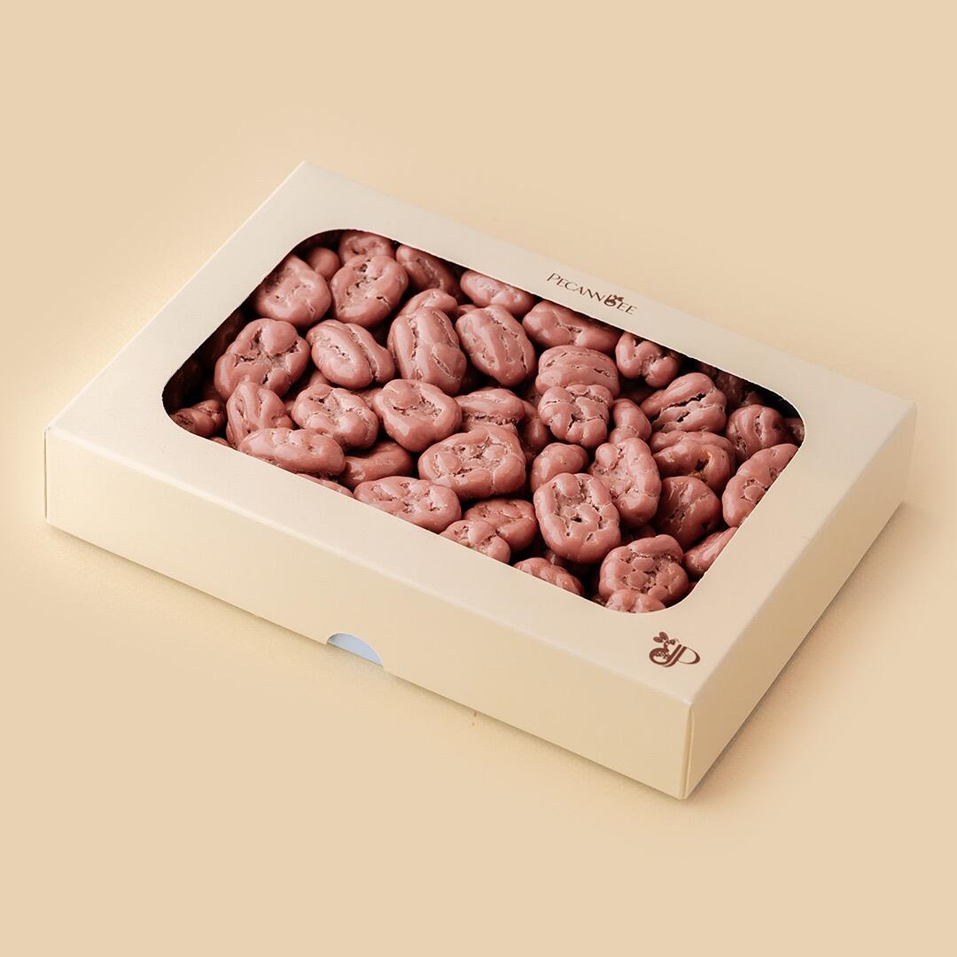 A high-angle studio shot of a rectangular cream-colored gift box with a transparent window filled with ruby chocolate-coated pecans by PecanBee on a beige backg