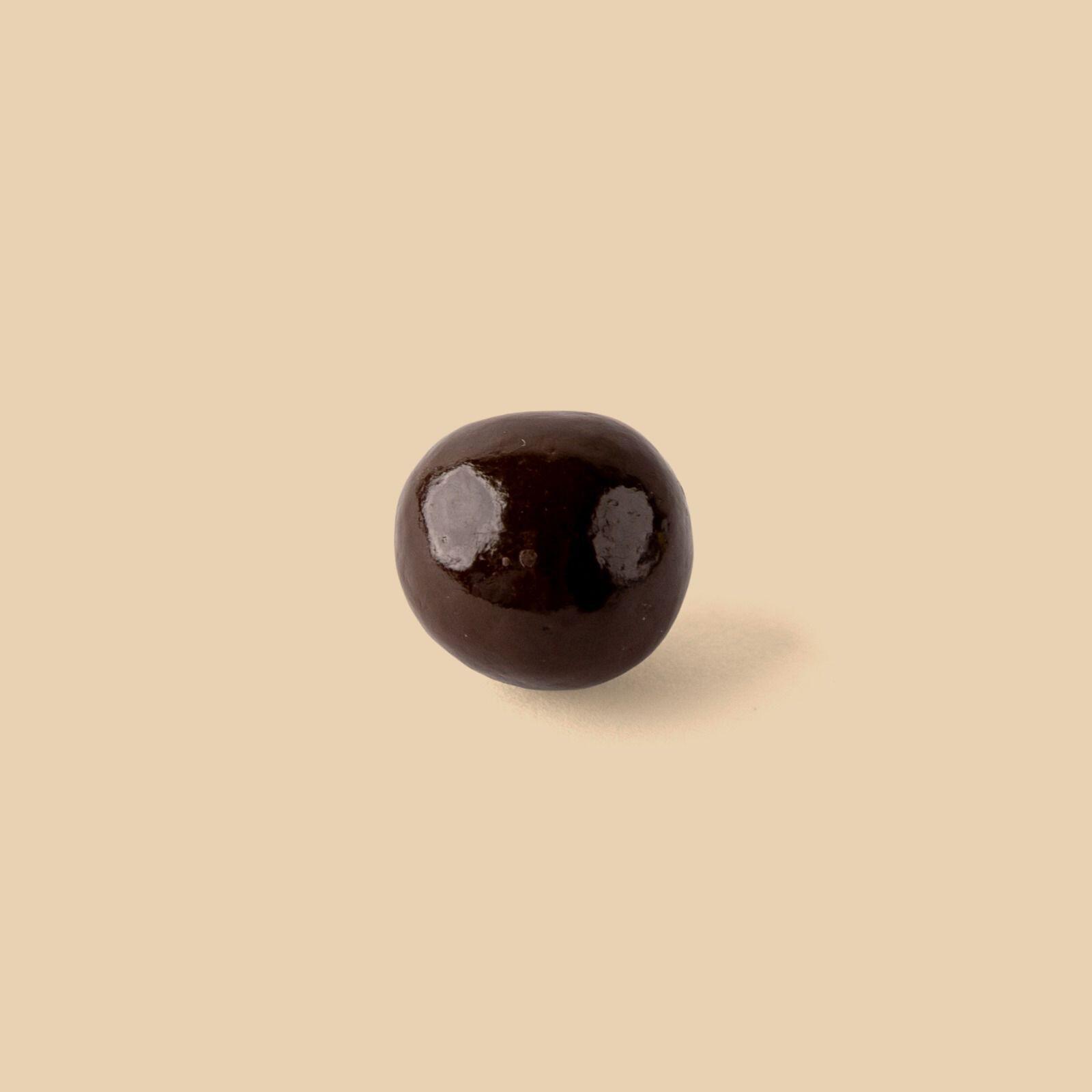 A single dark chocolate coated hazelnut dragee centered on a plain light beige background featuring a smooth glossy finish and a soft cast shadow