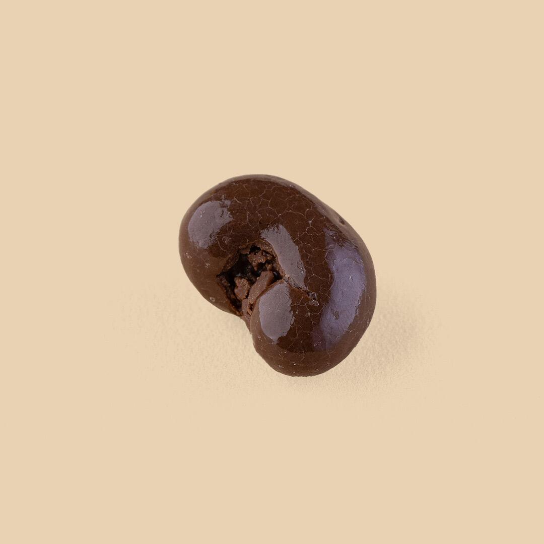 A single milk chocolate-coated cashew nut with a slightly cracked, glossy surface texture, centered on a solid light beige background.