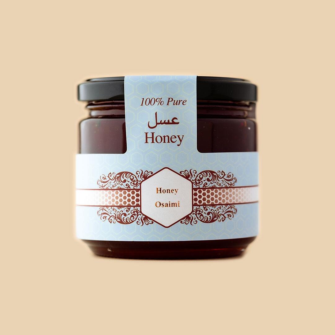 A glass jar of Osaimi honey with a black lid and a blue label featuring floral patterns, hexagonal honeycomb motifs, and the words 100% Pure Honey in English an