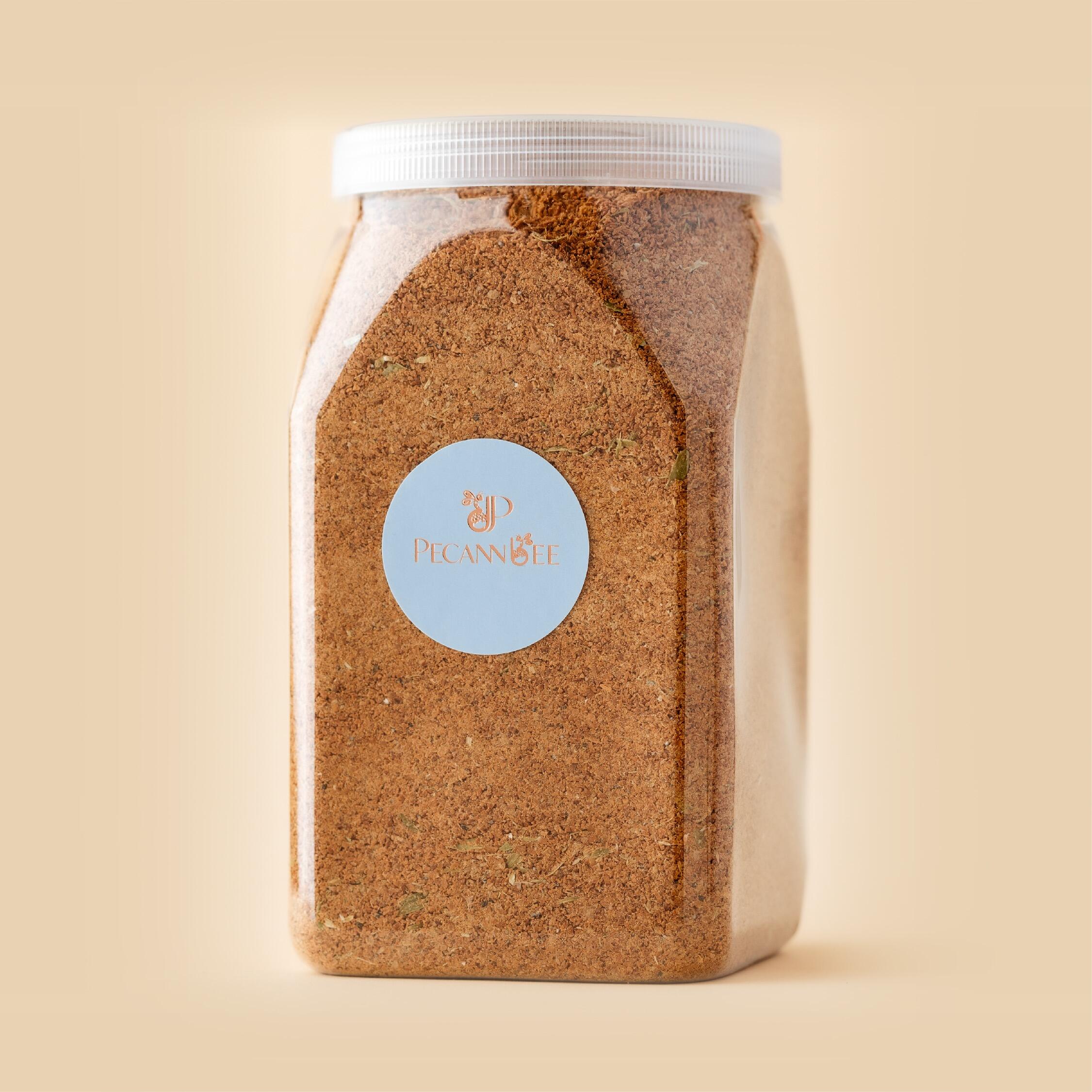 A clear, square plastic jar filled with fine, brown ground coffee, featuring a light blue PecanBee logo sticker, set against a smooth, minimalist light beige ba