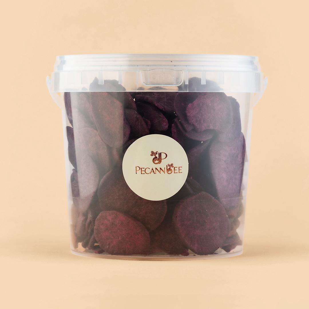 A clear, cylindrical plastic container filled with sliced, dehydrated purple sweet potato chips, centered against a solid, light beige background.