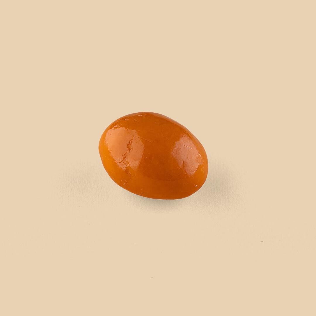 A single orange-glazed date candy centered against a solid light beige background, captured from a high-angle studio shot with soft lighting
