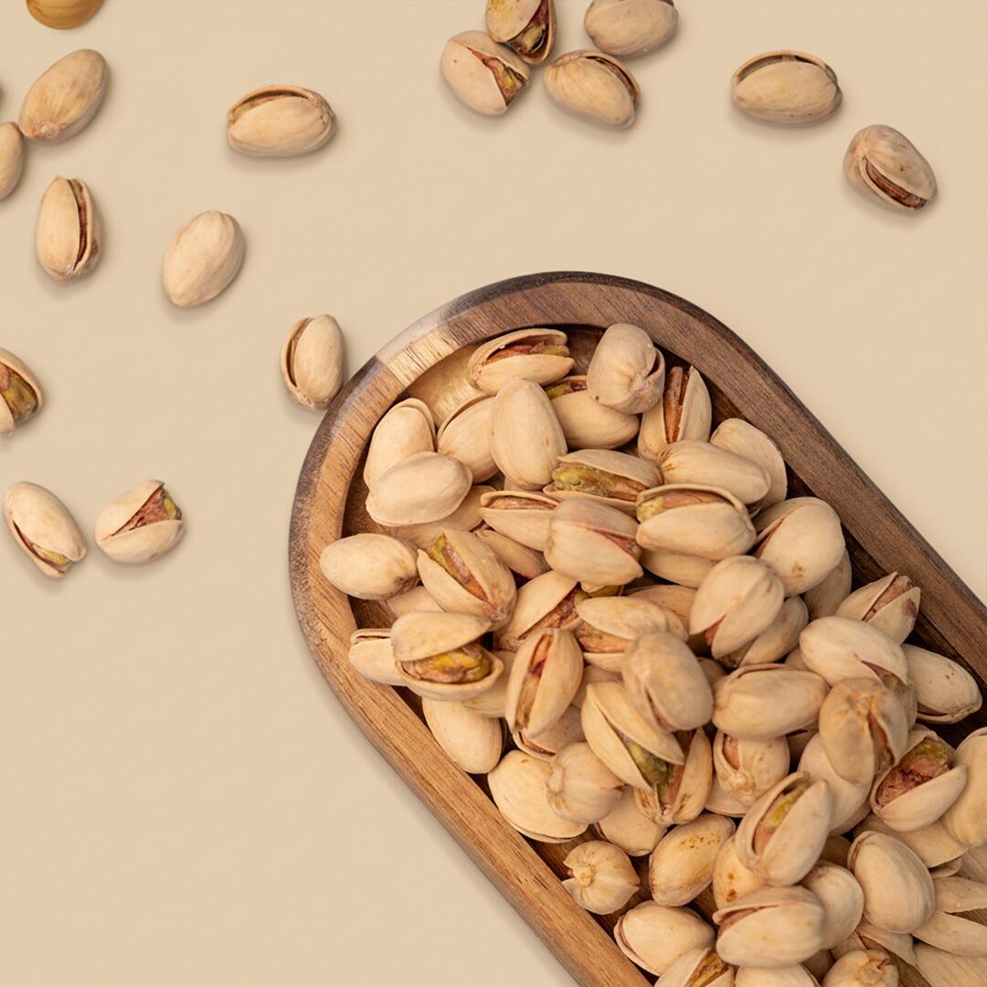 A rustic wooden bowl filled with salted Akbari pistachios, surrounded by scattered nuts on a neutral beige surface, shot from a top-down angle
