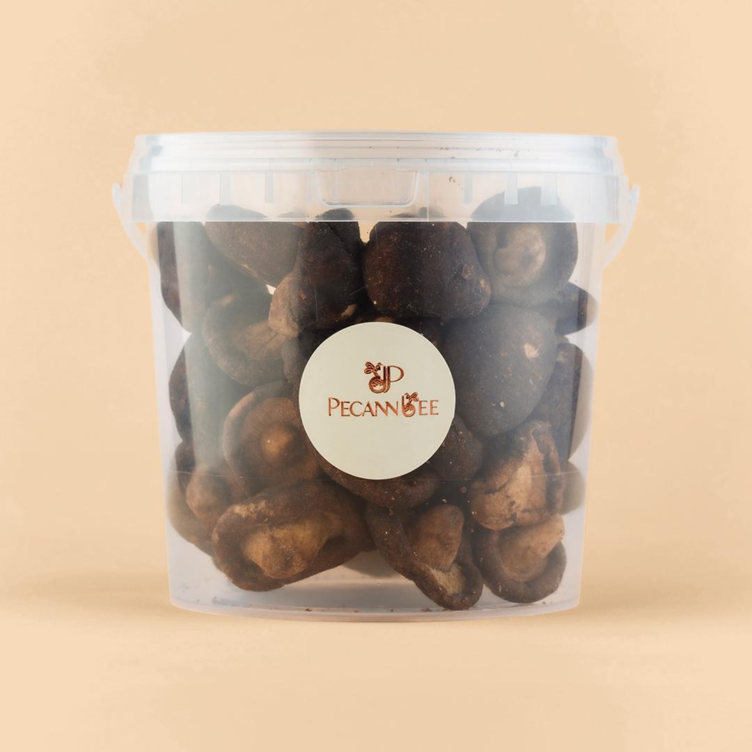 A clear plastic bucket filled with dark brown dehydrated mushrooms, centered with a circular PecanBee logo label, set against a solid beige background